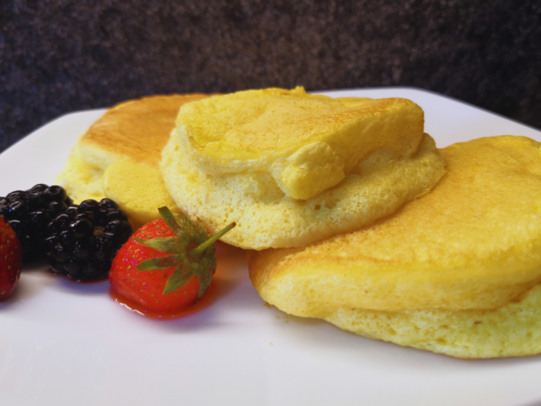 Soufflé pancake giapponesi alti e soffici, impilati su un piatto bianco, con sciroppo d'acero e frutti di bosco, fotografia food casalinga.